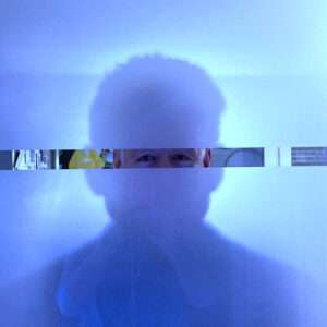 Eine künstlerische Porträtaufnahme in kühlen Blautönen. Die Silhouette eines Mannes ist fast vollständig hinter einer milchigen Glasfläche verborgen. Lediglich durch einen schmalen, klaren horizontalen Sehschlitz in Augenhöhe sind die wachen Augen direkt zu sehen. Das Bild spielt mit den Themen Sichtbarkeit, Identität und dem Wechselspiel zwischen Innen- und Außenansicht.