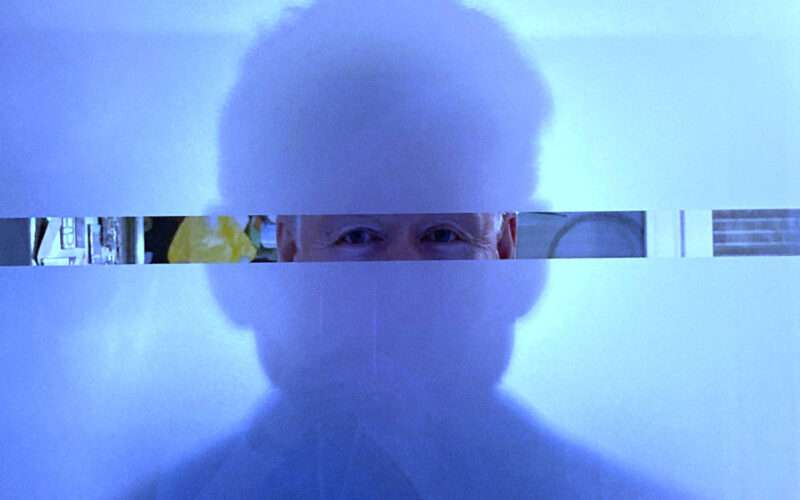 Eine künstlerische Porträtaufnahme in kühlen Blautönen. Die Silhouette eines Mannes ist fast vollständig hinter einer milchigen Glasfläche verborgen. Lediglich durch einen schmalen, klaren horizontalen Sehschlitz in Augenhöhe sind die wachen Augen direkt zu sehen. Das Bild spielt mit den Themen Sichtbarkeit, Identität und dem Wechselspiel zwischen Innen- und Außenansicht.